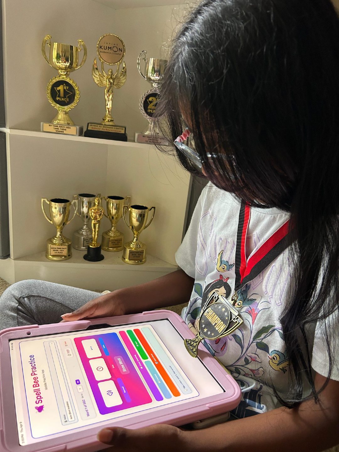 Young girl smiling with her spelling bee medal and trophies, holding a phone showing the Spell Bee Practice App